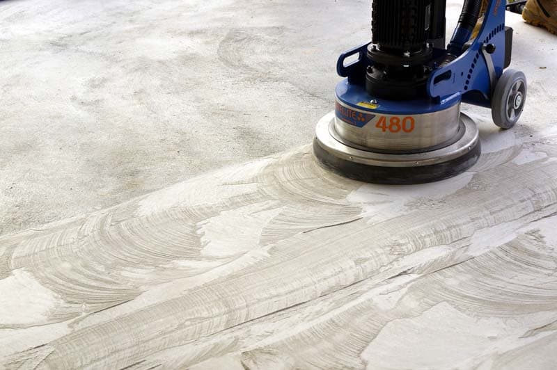 Blue industrial floor grinder smoothing a concrete surface, leaving circular patterns in the dust.